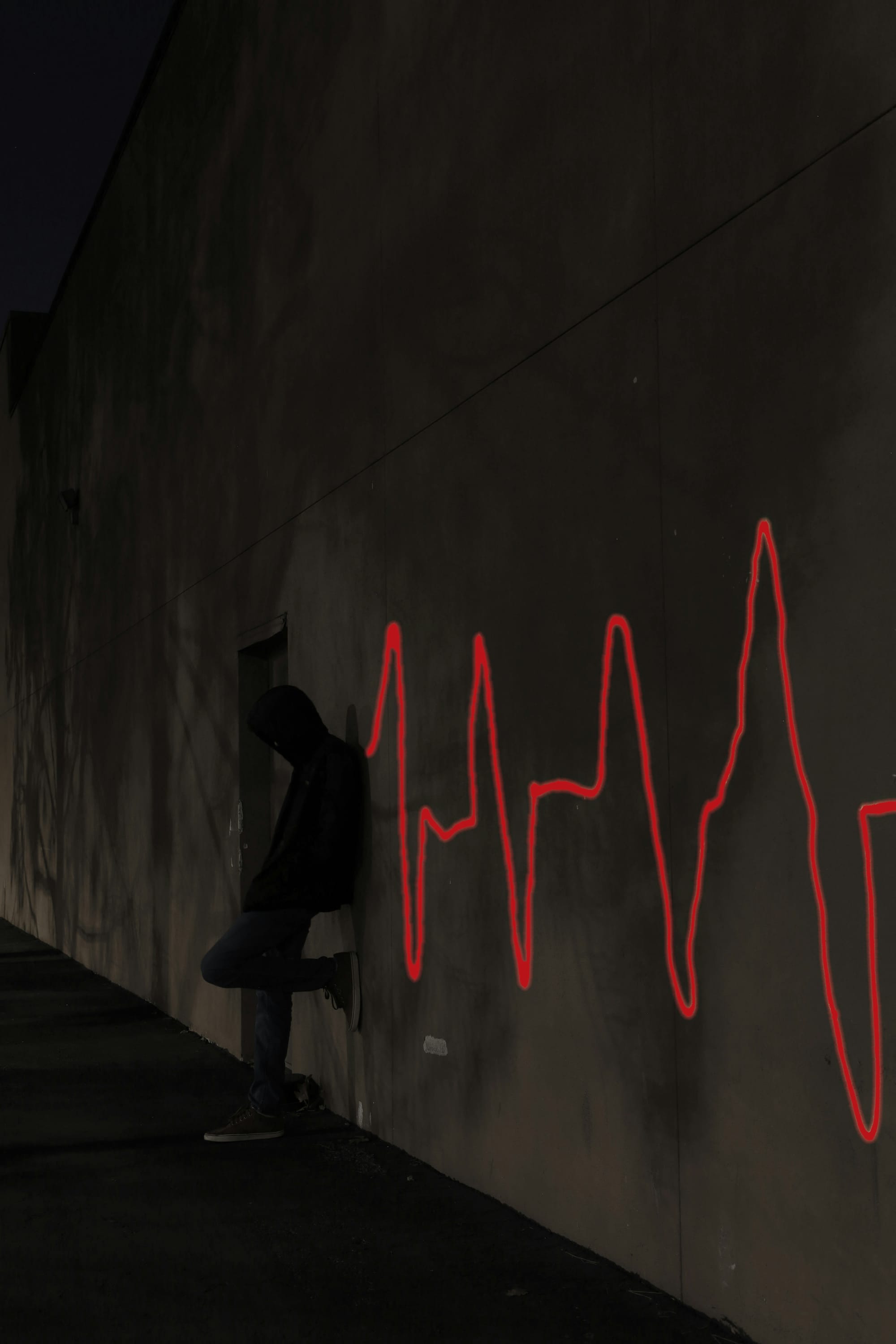 A hooded figure leans against a dark wall, in shadow, next to bright red graffiti in the shape of a pulsing heart monitor line stretches across the wall.